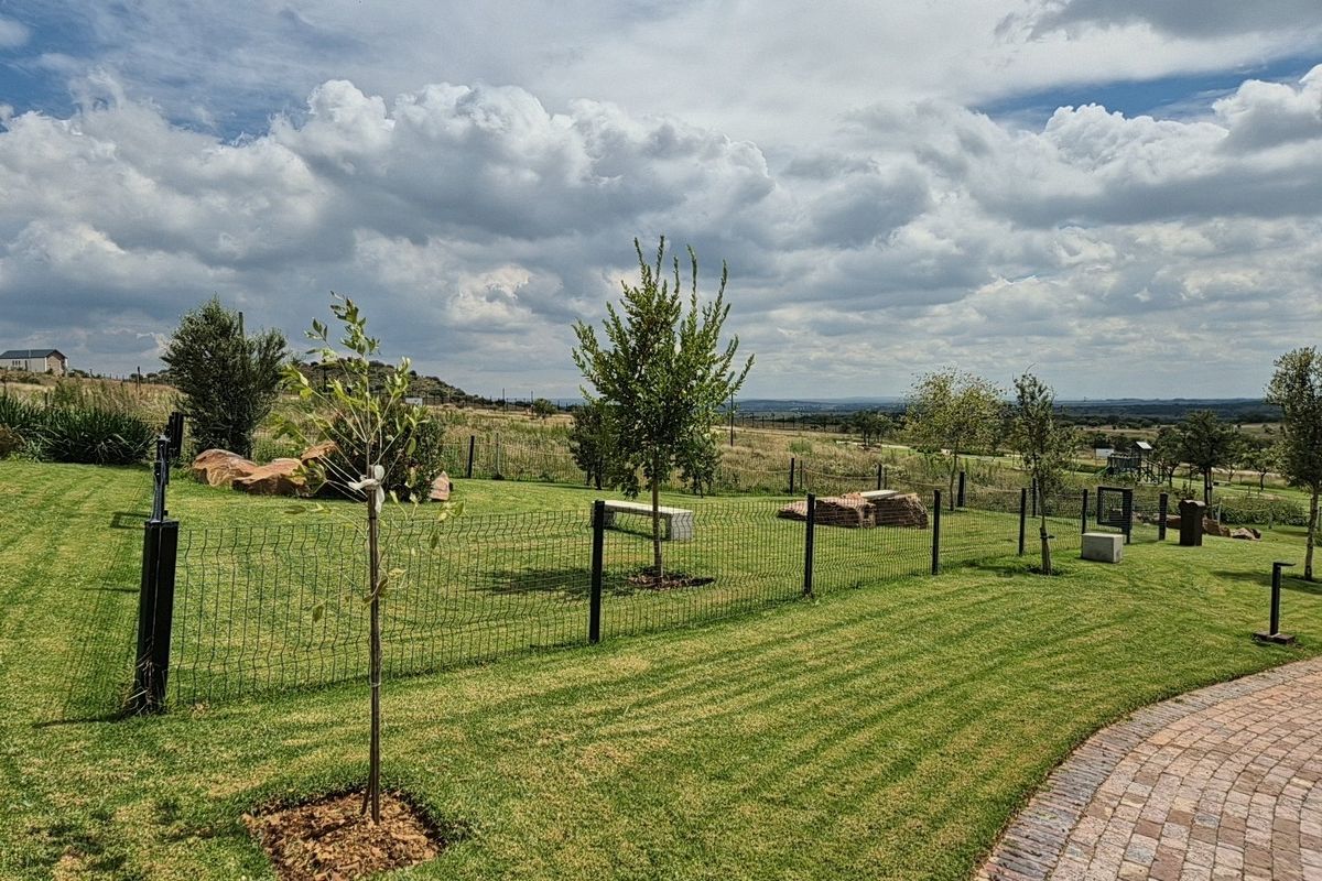 A well-maintained residential garden featuring a manicured lawn, young trees, wire mesh fencing, and a paved walkway with scenic landscape views.