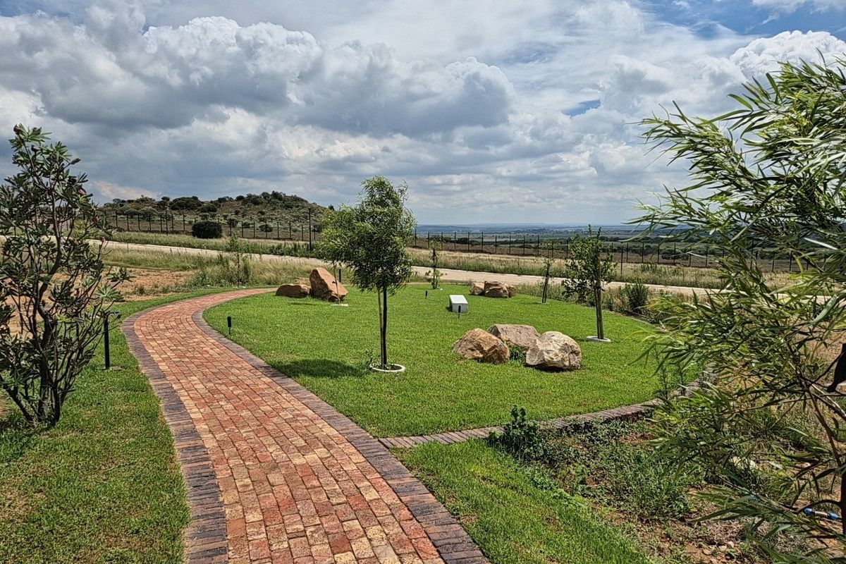 A well-maintained residential garden featuring a curved brick paved pathway, manicured lawn, decorative boulders, and scenic landscape views.