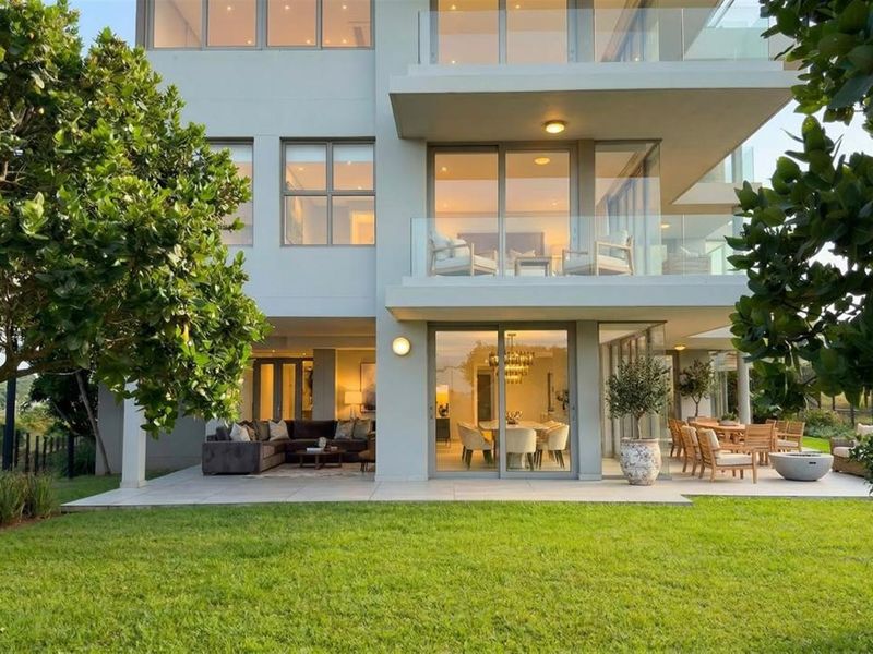 Rear view of a modern apartment building featuring a spacious paved patio, glass balustrades, and large sliding doors opening onto a well-maintained garden.