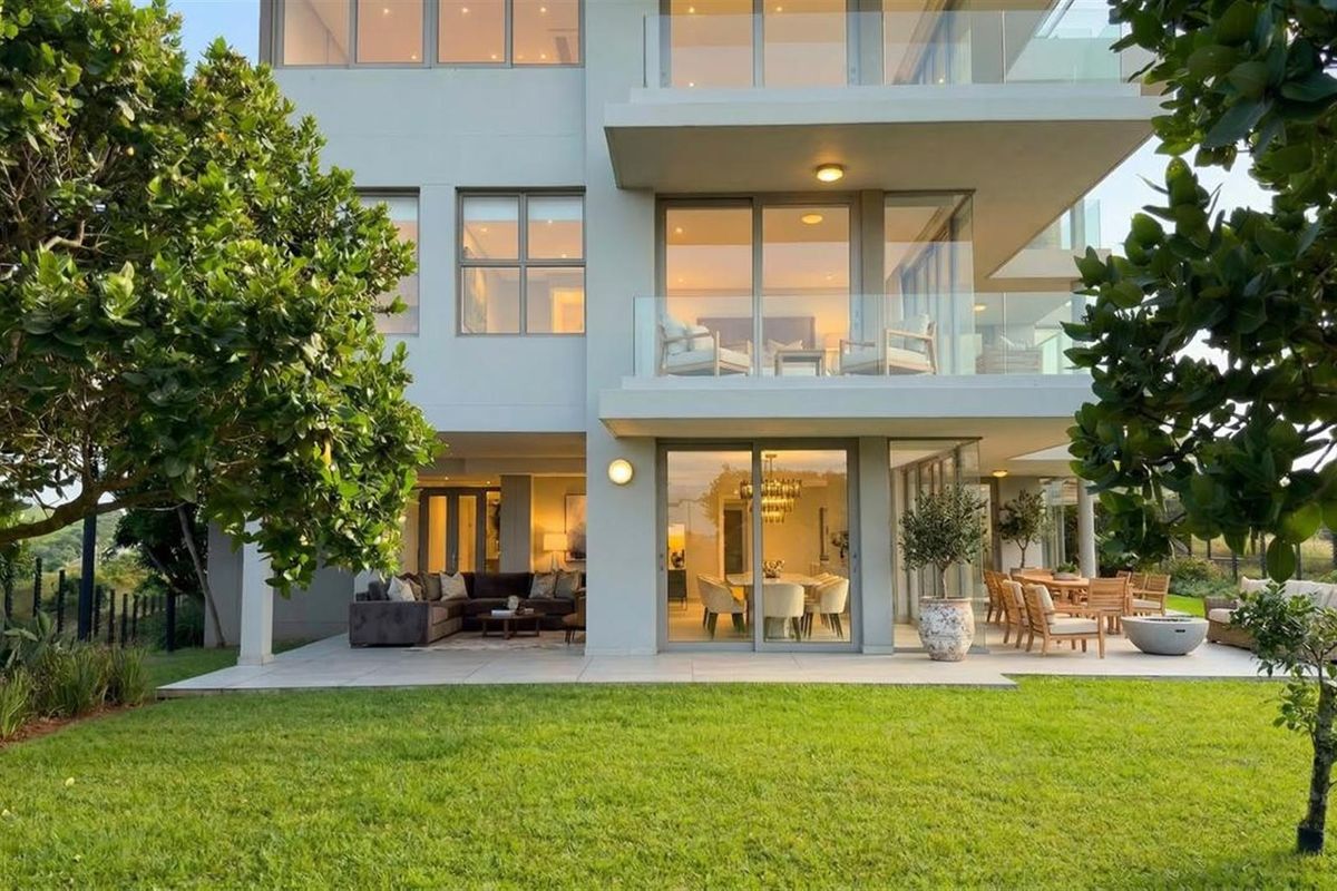 Rear view of a modern apartment building featuring a spacious paved patio, glass balustrades, and large sliding doors opening onto a well-maintained garden.
