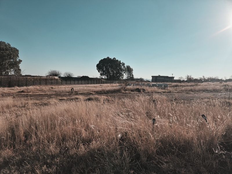 A wide-angle view of a vacant residential plot featuring dry grass, perimeter fencing, and distant structures under a clear sky.