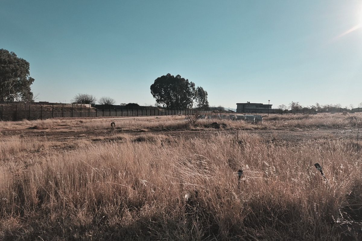 A wide-angle view of a vacant residential plot featuring dry grass, perimeter fencing, and distant structures under a clear sky.