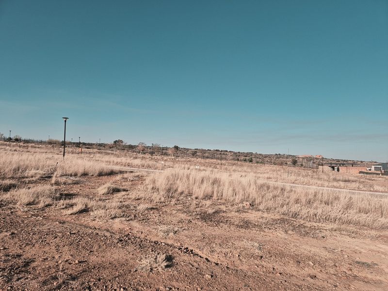 A view of a vacant residential plot featuring natural grass, perimeter fencing, and street lighting infrastructure.