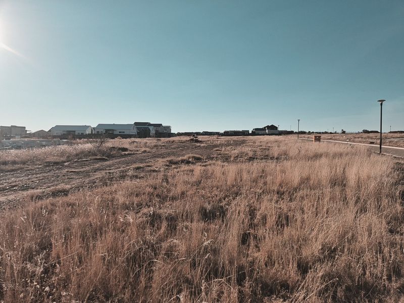 A vacant residential plot featuring dry grass, a paved pathway, and street lighting, situated in a developing neighborhood.