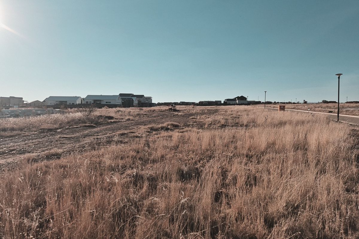 A vacant residential plot featuring dry grass, a paved pathway, and street lighting, situated in a developing neighborhood.