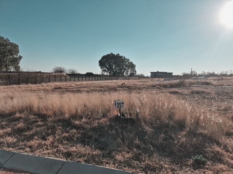 A vacant residential plot featuring dry grass, perimeter fencing, and clear street-level access, suitable for new development.