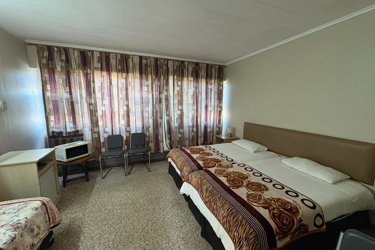 A dated hotel bedroom featuring wall-to-wall carpeting, a built-in headboard, and large window curtains, offering a functional space in need of modernization.
