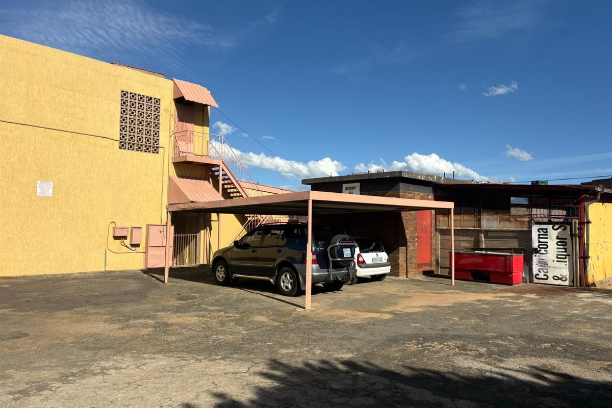 Rear exterior view of the hotel property featuring a covered carport, external staircase, and paved parking area with weathered building finishes.