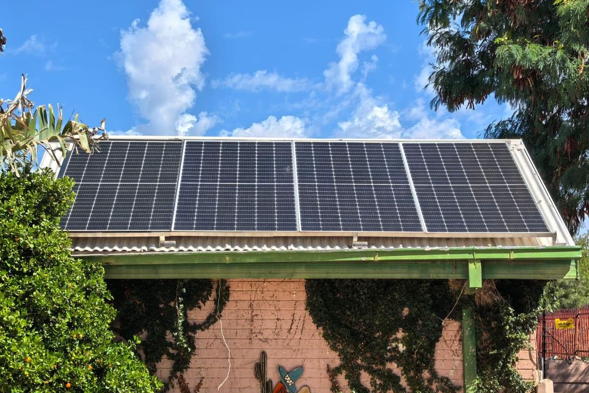 Exterior view of a residential roof featuring a modern solar panel array mounted on corrugated roofing above a brick wall with climbing vegetation.