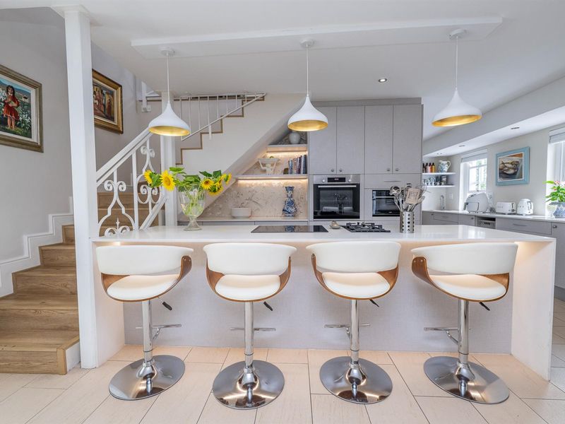 Modern open-plan kitchen featuring a central island with breakfast bar, integrated appliances, stone countertops, and an adjacent staircase with decorative railing.