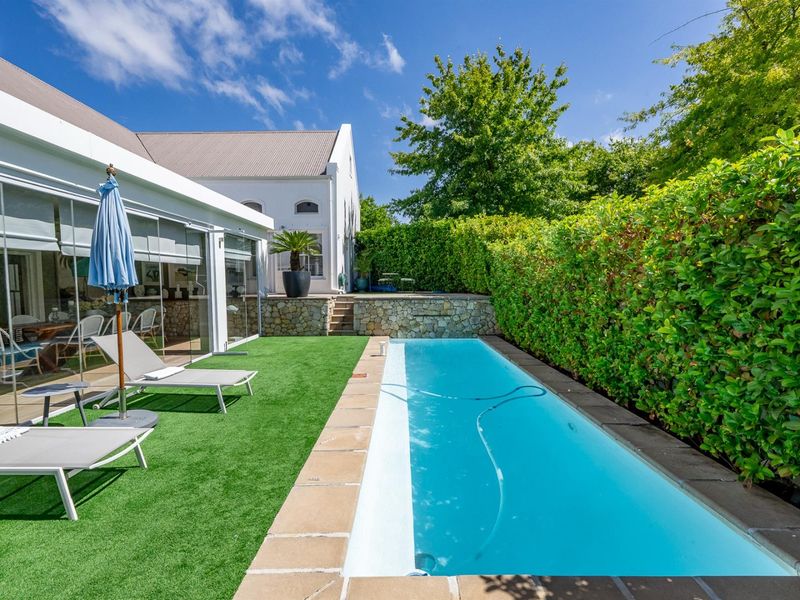 A well-maintained outdoor pool area featuring a rectangular swimming pool, stone retaining wall, and artificial turf, adjacent to a glass-enclosed patio.