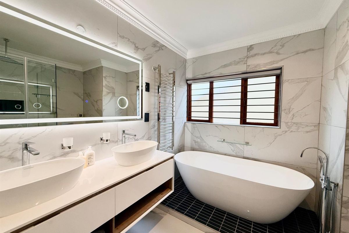 Modern, high-end bathroom featuring a double vanity with backlit mirror, a sleek freestanding bathtub, and elegant marble-look wall tiling.