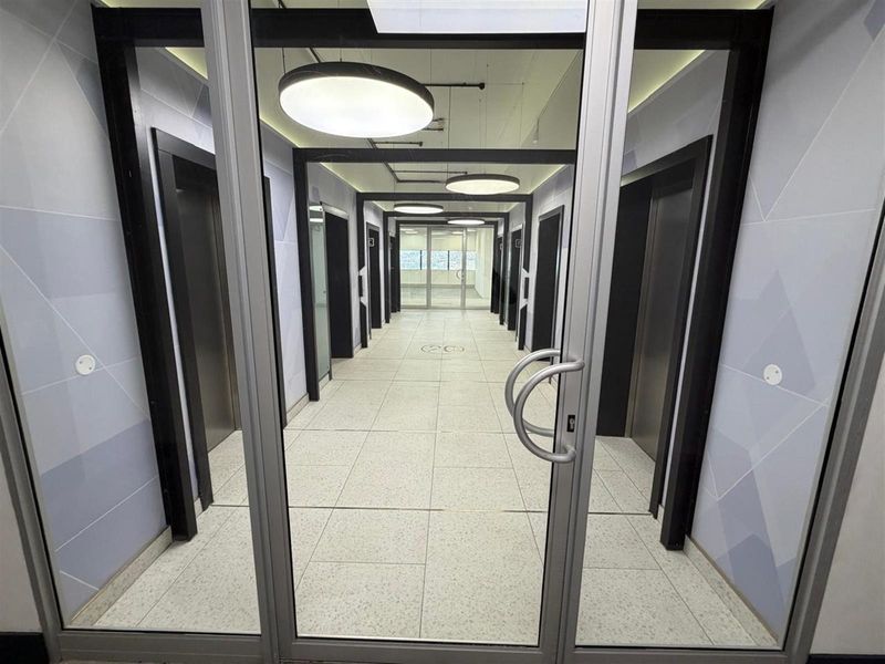 A modern office building hallway featuring tiled flooring, multiple elevator access points, and contemporary recessed ceiling lighting.