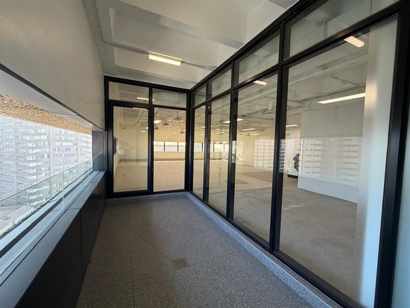 A modern, covered balcony featuring terrazzo flooring, a sleek glass balustrade, and expansive black-framed glass partitions overlooking the office interior.