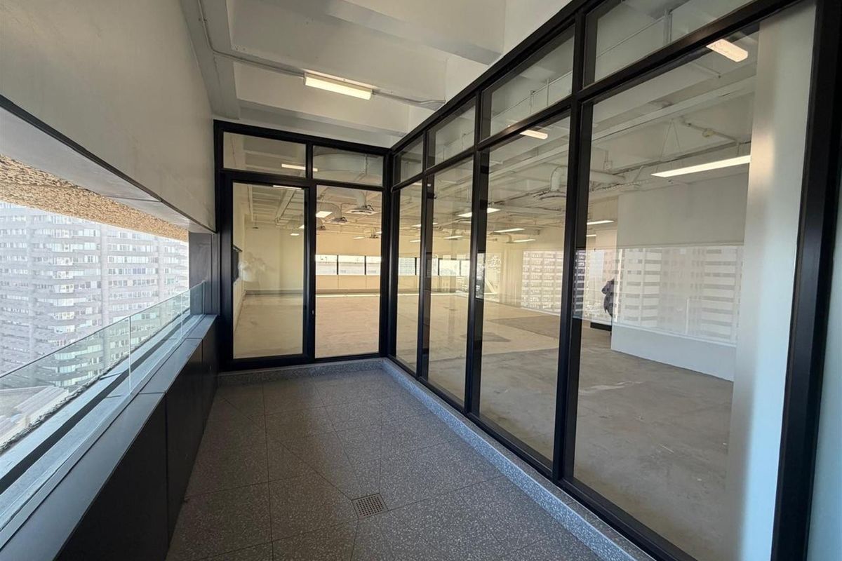 A modern, covered balcony featuring terrazzo flooring, a sleek glass balustrade, and expansive black-framed glass partitions overlooking the office interior.