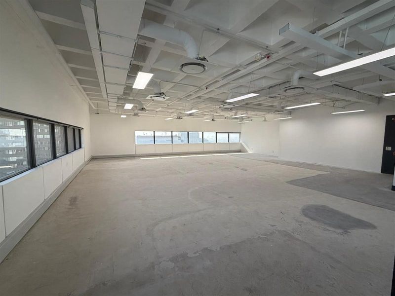 A spacious, open-plan office area featuring an industrial-style exposed ceiling with ductwork, linear lighting, and large windows providing ample natural light.