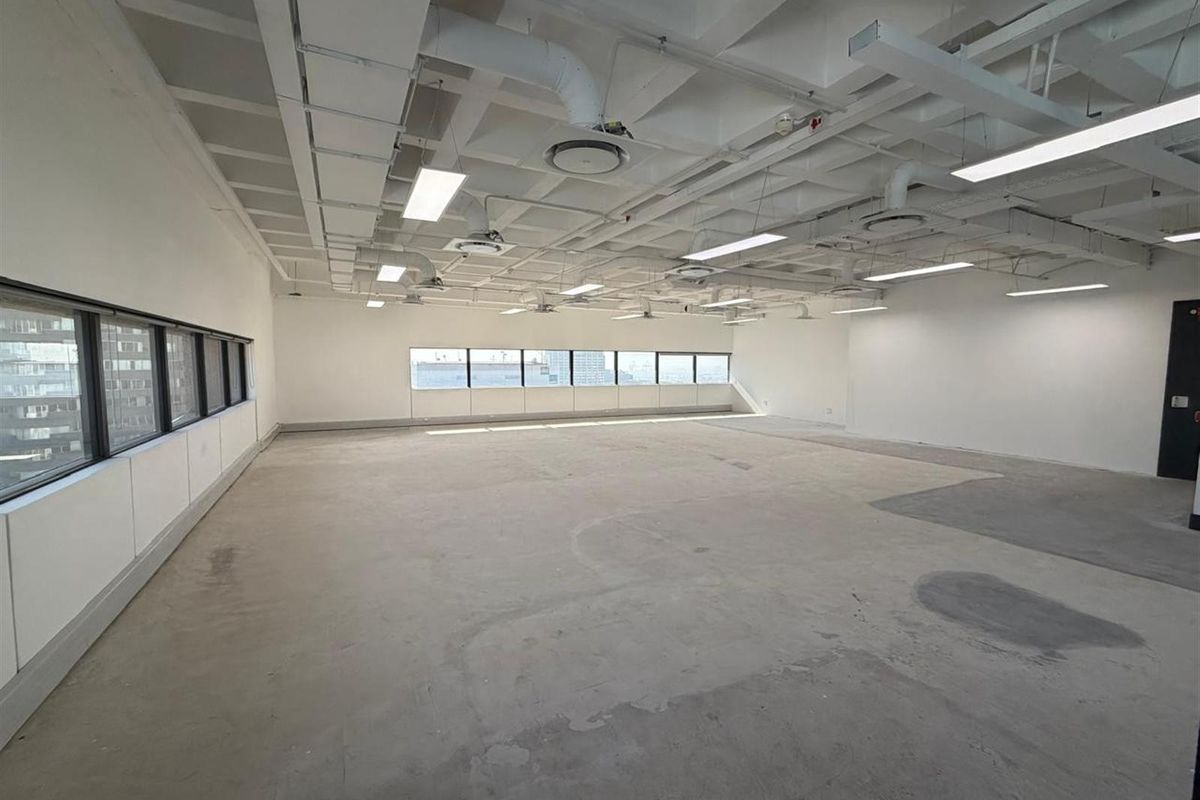 A spacious, open-plan office area featuring an industrial-style exposed ceiling with ductwork, linear lighting, and large windows providing ample natural light.