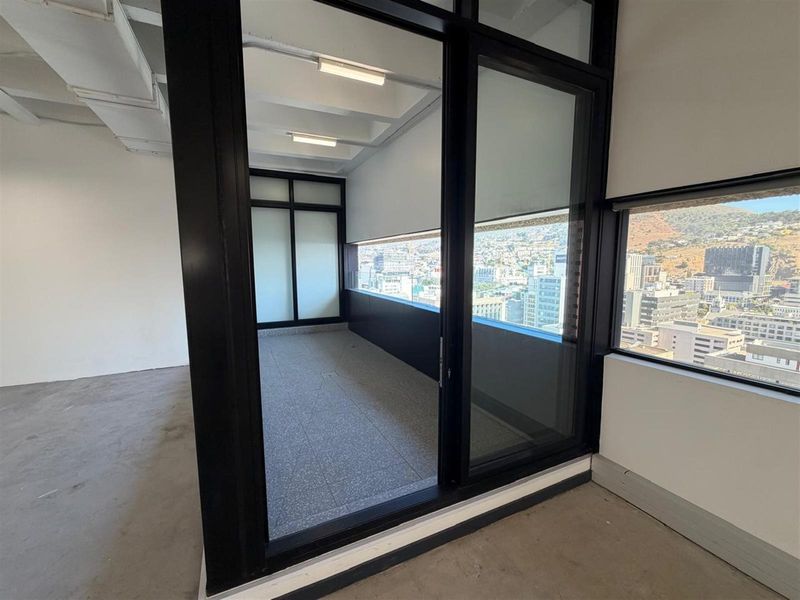 Modern empty office space featuring exposed ceiling beams, terrazzo flooring, and large black-framed glass doors opening to a balcony with city views.