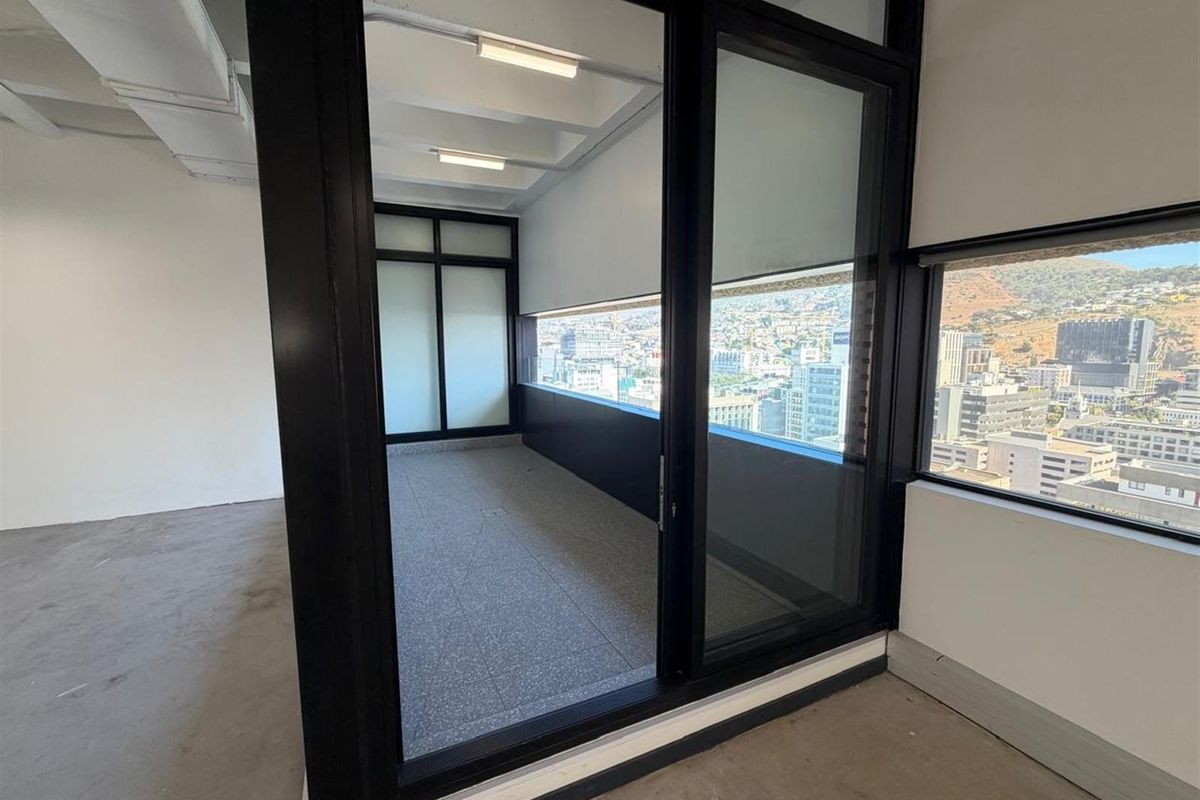 Modern empty office space featuring exposed ceiling beams, terrazzo flooring, and large black-framed glass doors opening to a balcony with city views.