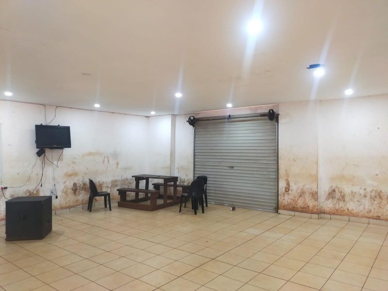 Open plan commercial space featuring tiled flooring, recessed lighting, and a secure roller shutter door, currently requiring significant wall repairs and cosmetic refurbishment.