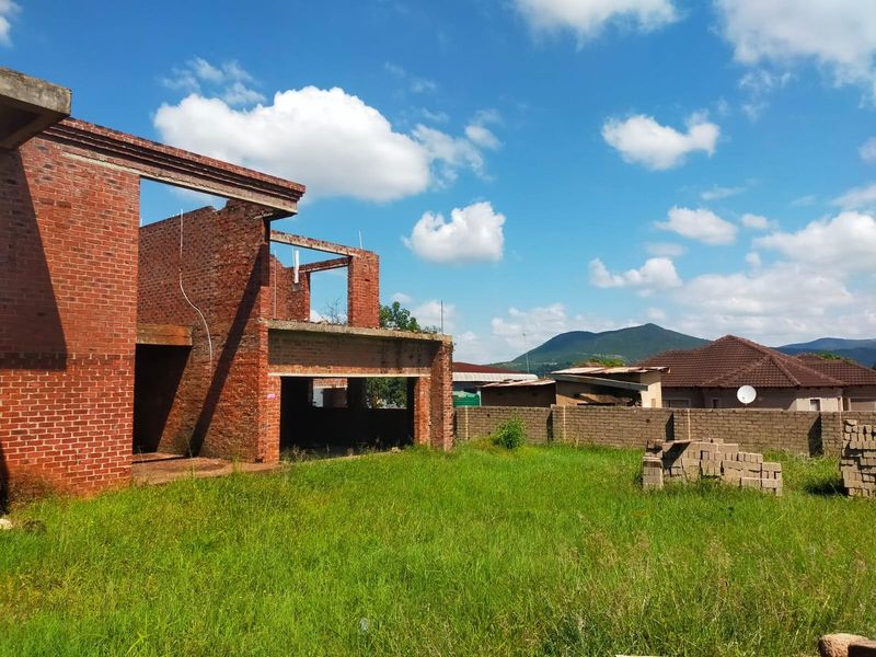 A view of an incomplete residential property under construction, featuring exposed brickwork and structural concrete elements set within a large grassed yard.