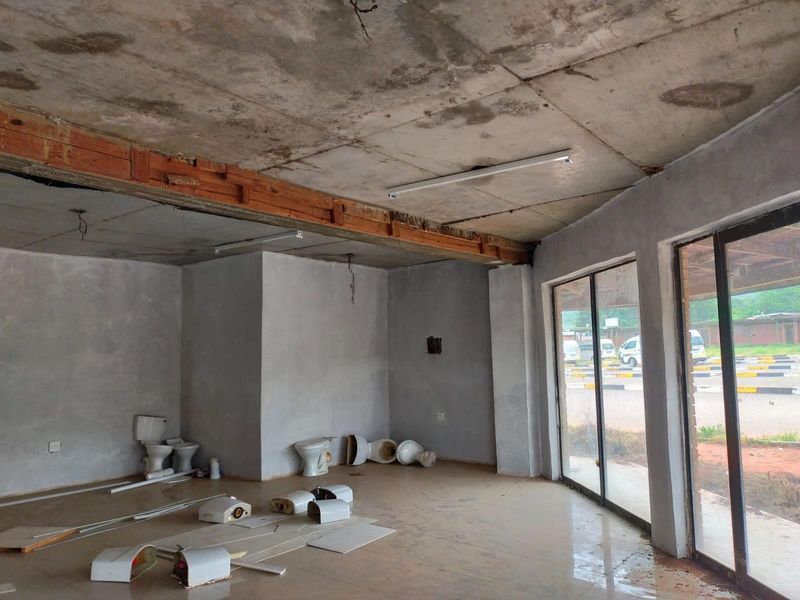An unfinished commercial space featuring exposed concrete ceilings, large glass storefront windows, and tiled flooring, currently in a shell condition.