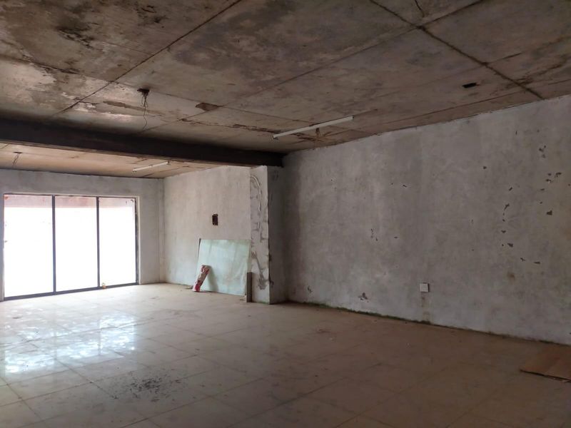 An empty, open-plan commercial space featuring tiled flooring, an exposed concrete ceiling, and large sliding glass doors, currently requiring significant renovation and cosmetic repairs.