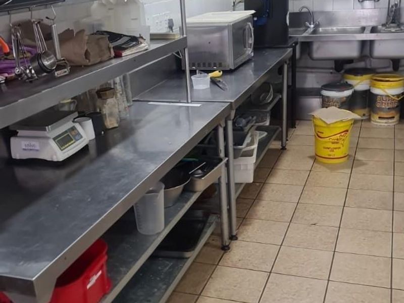 A functional commercial kitchen featuring stainless steel prep stations, a double bowl sink, wall-mounted shelving, and durable tiled surfaces throughout.