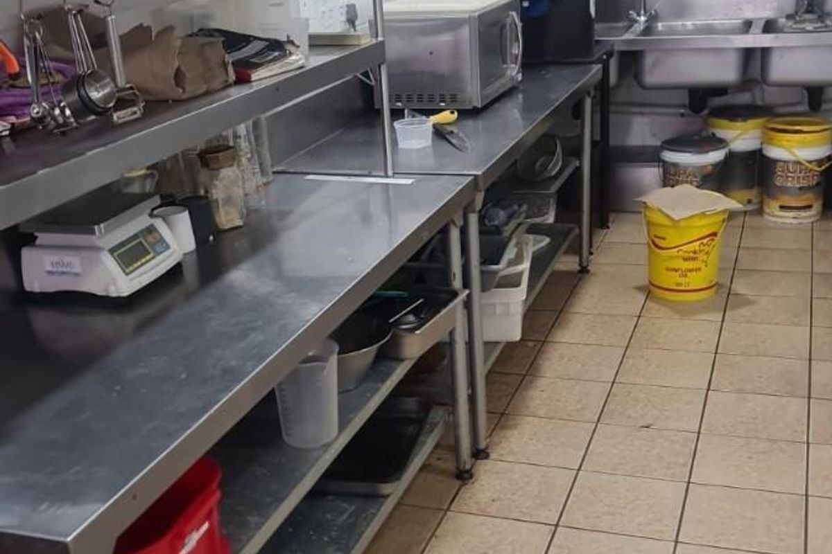 A functional commercial kitchen featuring stainless steel prep stations, a double bowl sink, wall-mounted shelving, and durable tiled surfaces throughout.