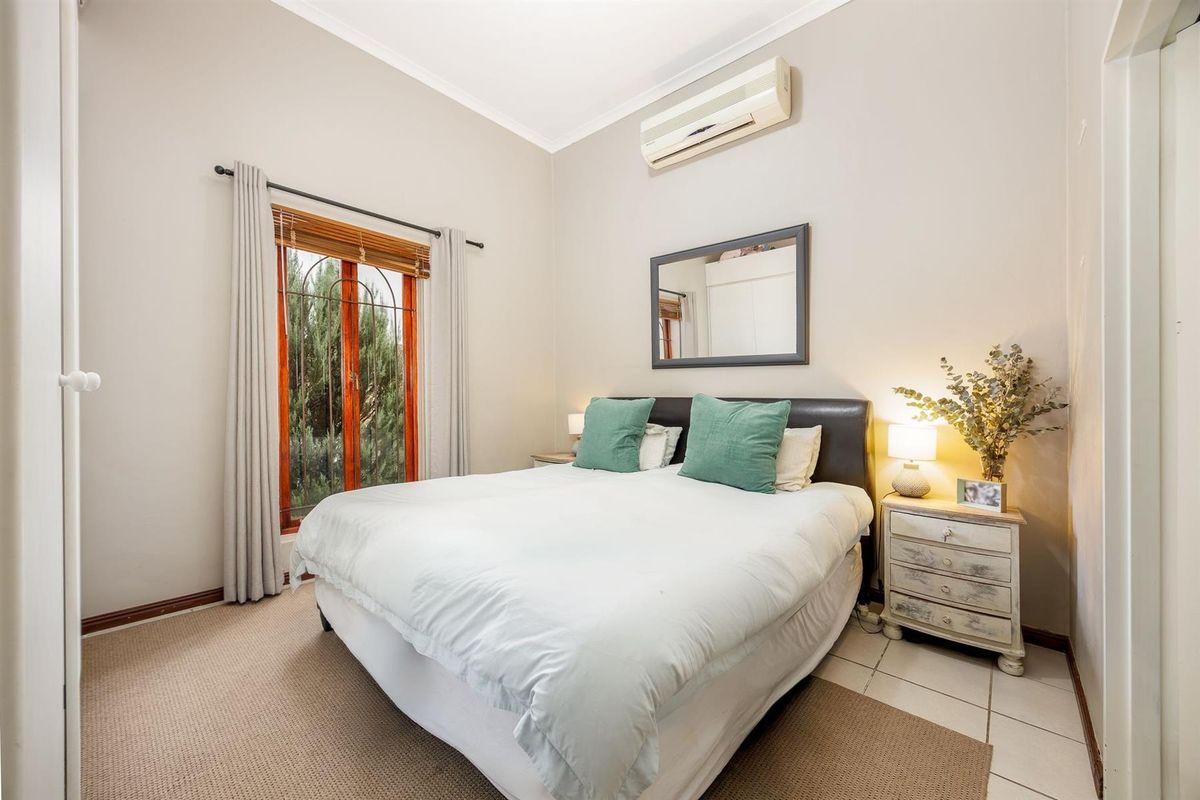 A cozy bedroom featuring a wall-mounted air conditioning unit, tiled flooring, and a window with security bars overlooking the garden.