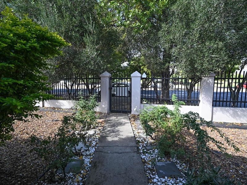 A gated pedestrian entrance featuring a concrete pathway leading to a metal security gate, framed by established trees and a perimeter fence.