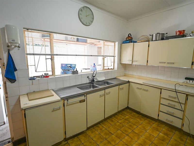 A functional kitchen featuring dated cabinetry, a double stainless steel sink, tiled splashbacks, and original floor tiles with a large window overlooking the exterior.