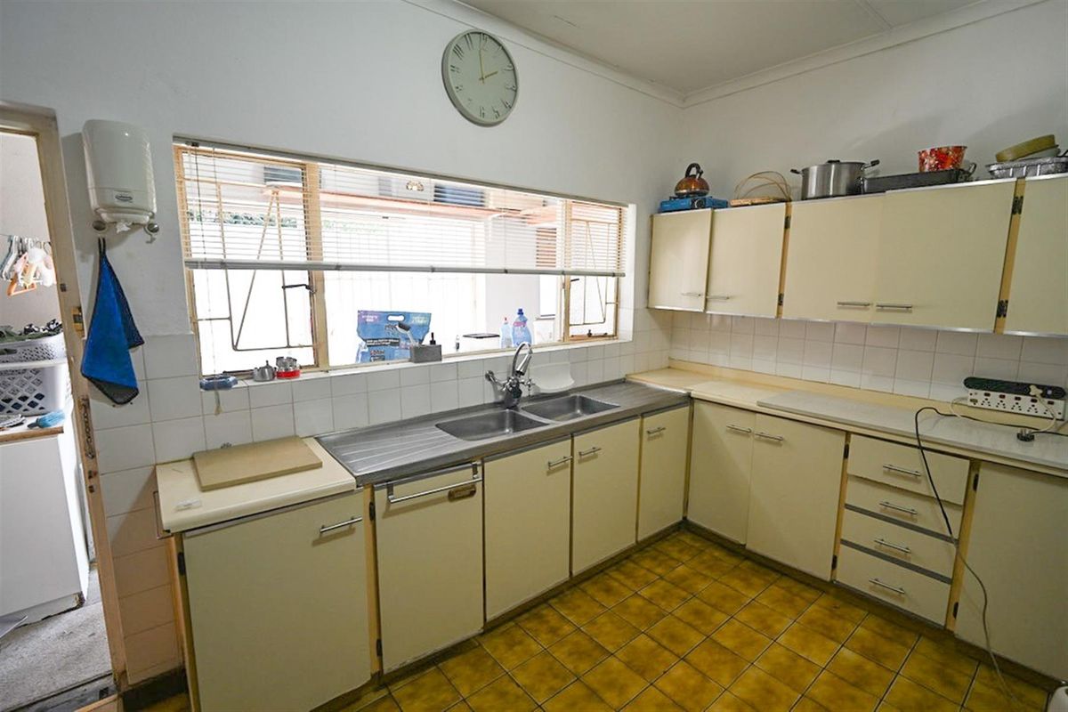 A functional kitchen featuring dated cabinetry, a double stainless steel sink, tiled splashbacks, and original floor tiles with a large window overlooking the exterior.