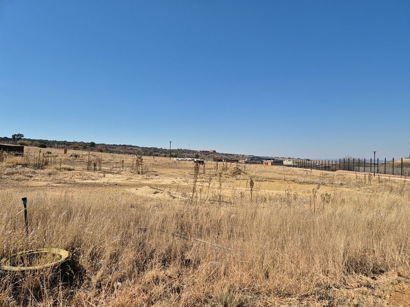 A view of a vacant residential plot featuring dry grass, perimeter fencing, and an existing utility connection point.