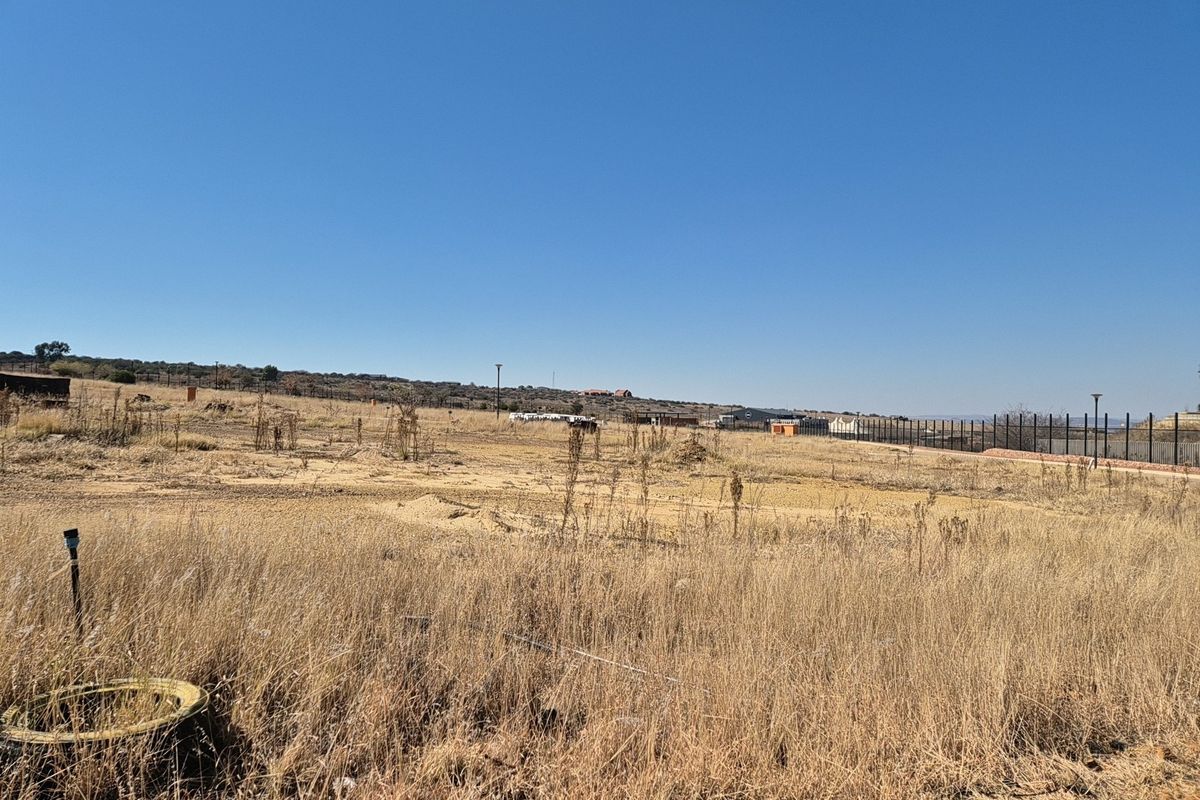 A view of a vacant residential plot featuring dry grass, perimeter fencing, and an existing utility connection point.