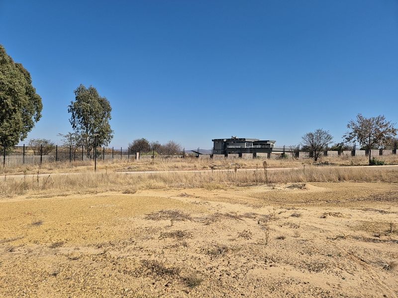 A view of a vacant residential plot featuring dry vegetation, perimeter fencing, and an unpaved access road.