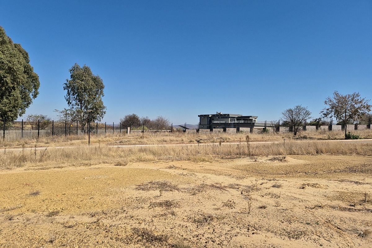 A view of a vacant residential plot featuring dry vegetation, perimeter fencing, and an unpaved access road.