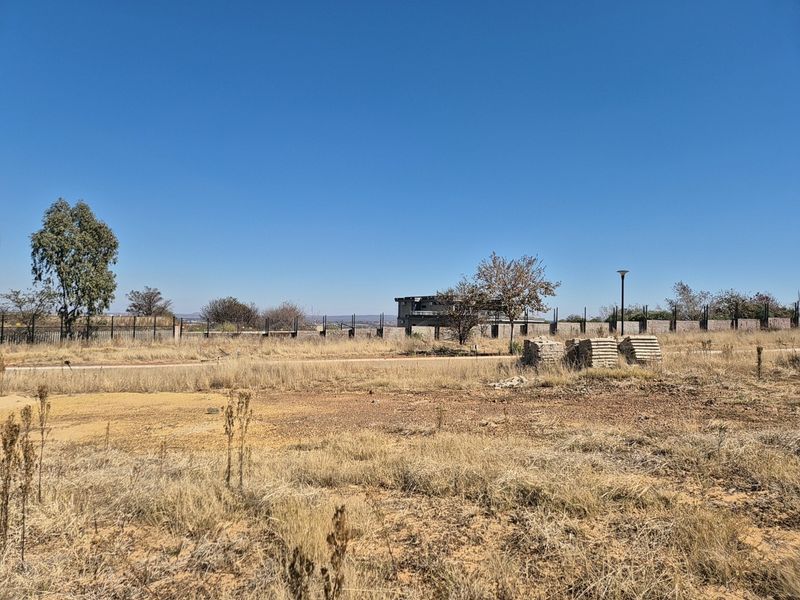 A vacant residential plot featuring dry grass, perimeter fencing, and distant views of neighboring structures under a clear blue sky.