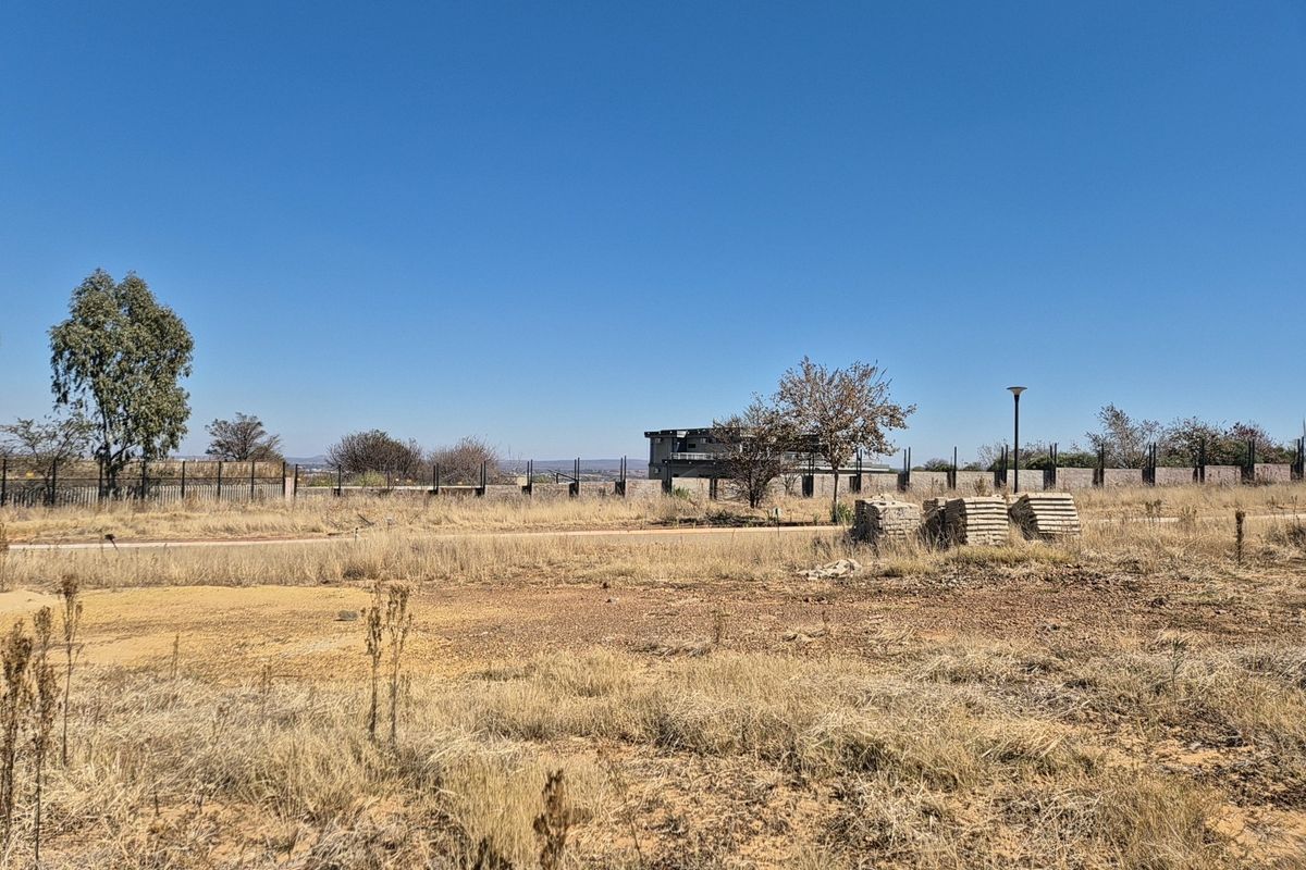 A vacant residential plot featuring dry grass, perimeter fencing, and distant views of neighboring structures under a clear blue sky.