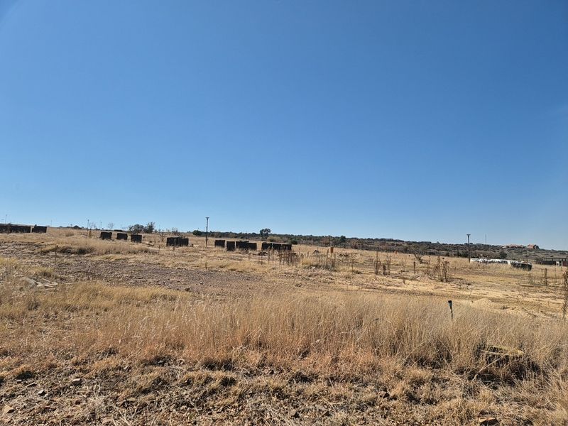 A wide-angle view of a vacant residential plot featuring dry grass, perimeter fencing, and utility poles under a clear blue sky.