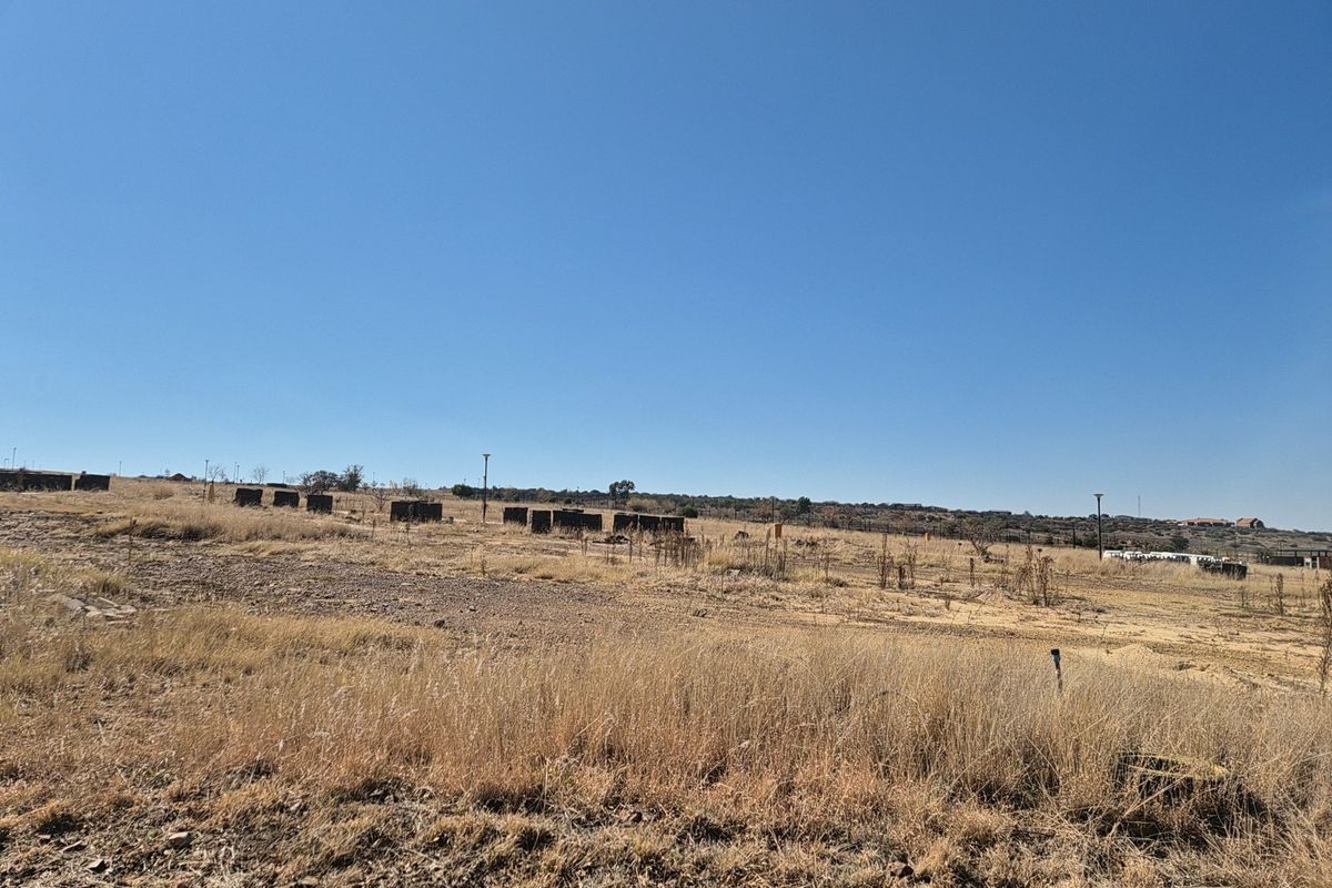 A wide-angle view of a vacant residential plot featuring dry grass, perimeter fencing, and utility poles under a clear blue sky.