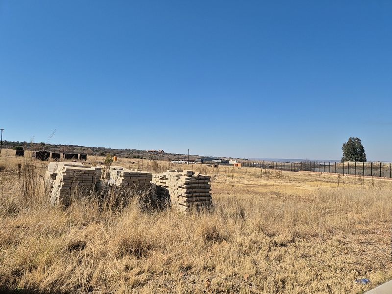 A vacant residential plot featuring dry grass, perimeter fencing, and stacks of building materials, offering a clear view of the surrounding landscape.