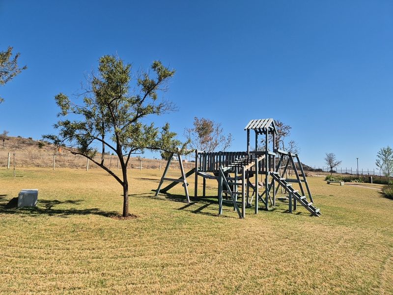 A spacious residential garden featuring a wooden playground structure set within a well-maintained lawn area under a clear blue sky.