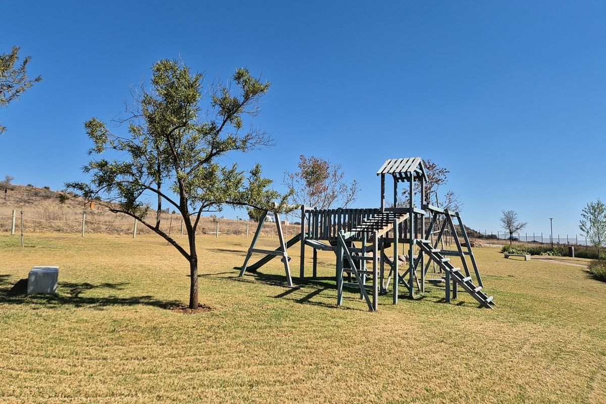 A spacious residential garden featuring a wooden playground structure set within a well-maintained lawn area under a clear blue sky.