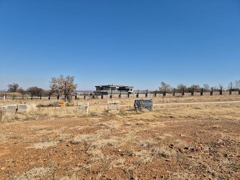 A view of a vacant residential plot featuring dry grass, perimeter fencing, and a distant view of neighboring properties.