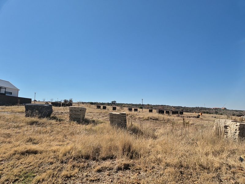 A wide-angle view of a vacant residential plot featuring dry grass and scattered construction materials under a clear blue sky.