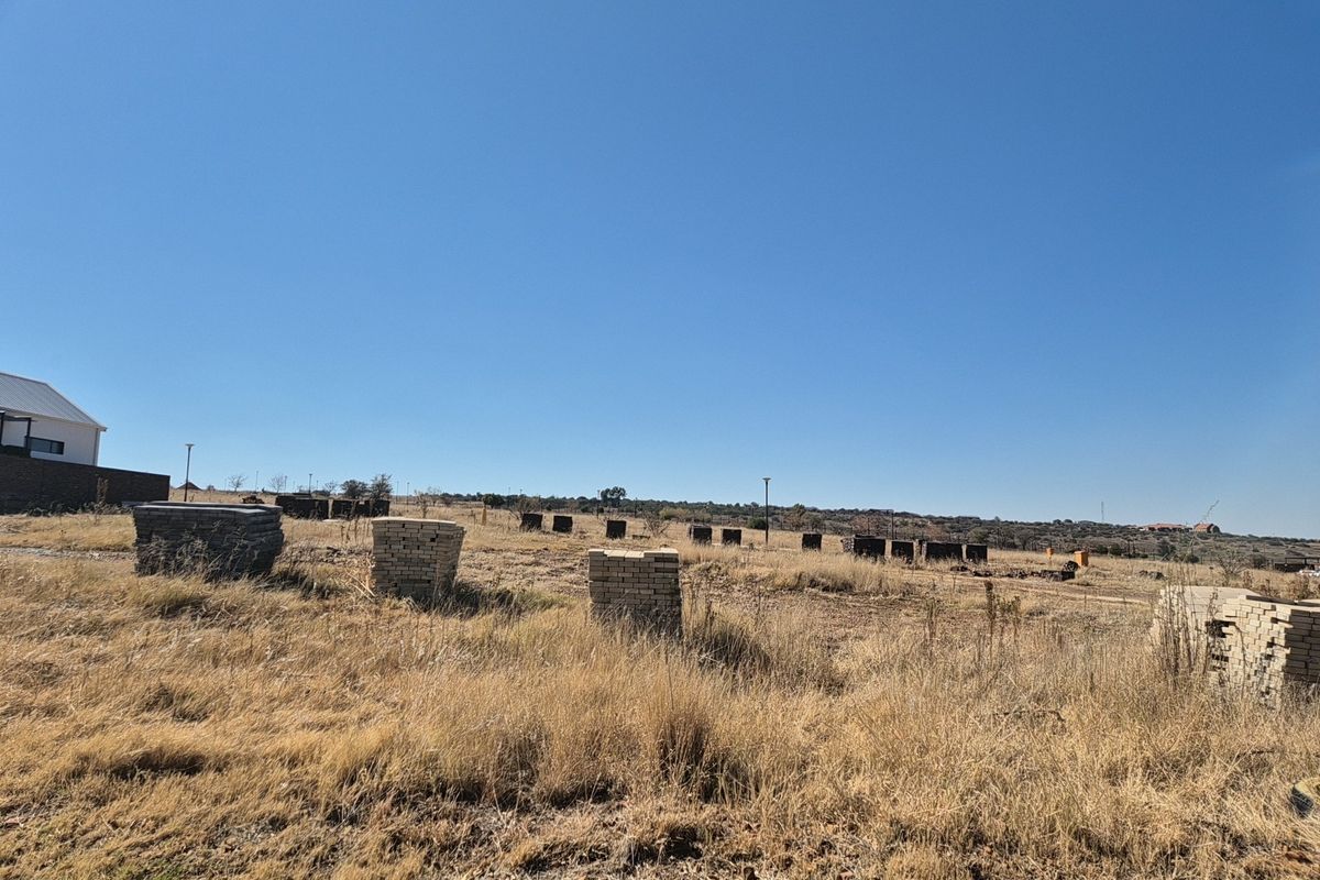 A wide-angle view of a vacant residential plot featuring dry grass and scattered construction materials under a clear blue sky.