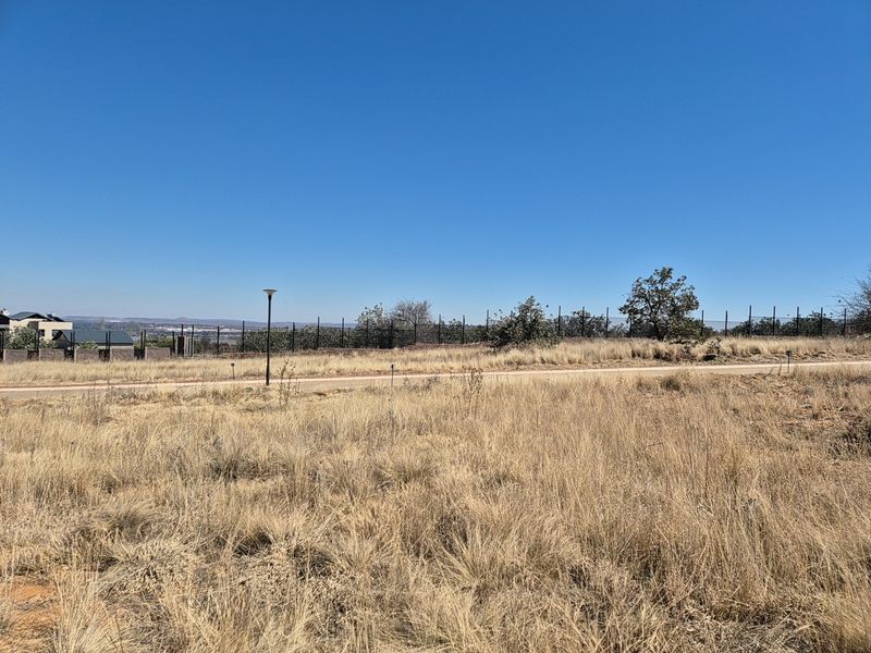 A vacant residential plot featuring dry grass, perimeter fencing, and street lighting, situated in a developed estate setting.
