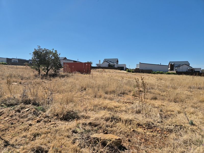 A view of a vacant residential plot featuring dry grass and surrounding neighborhood homes in the background.