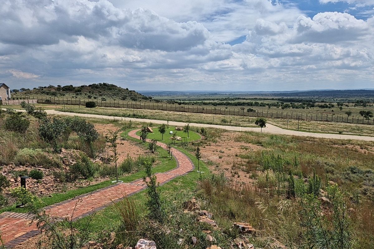 A scenic view of the residential land featuring a brick paved pathway, landscaped garden areas, and expansive open landscape with perimeter fencing.